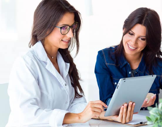 Woman wearing a white coat holding a tablet discussing healthcare management technology with the woman in blue